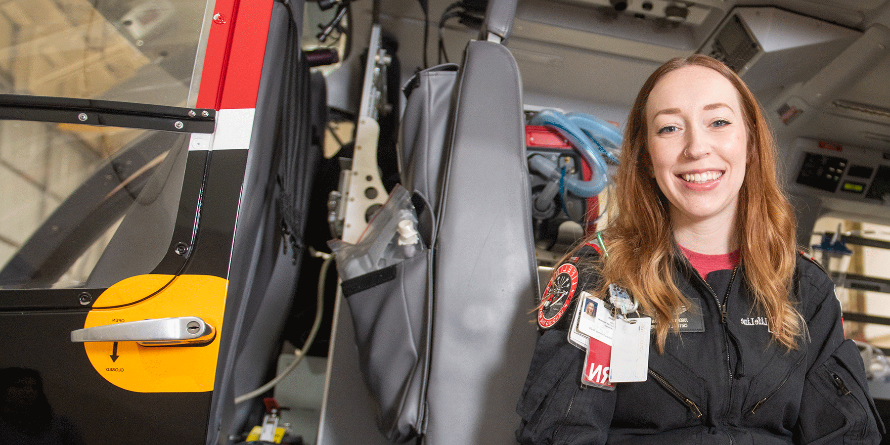 A white female student poses in an emergency services LifeLine vehicle. She has long red hair, a nose ring, and she wears a back LifeLine uniform with a red undershirt. A badge is on her chest. Medical devices and equipment are in the vehicle.