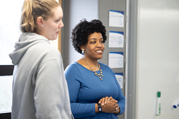 Two women standing in a classroom or office setting, engaged in conversation. The woman on the left, wearing a blue dress and a necklace, smiles warmly, while the woman on the right, dressed casually in a gray hoodie, listens attentively. A whiteboard with notes or signs is visible in the background, suggesting an educational or professional environment