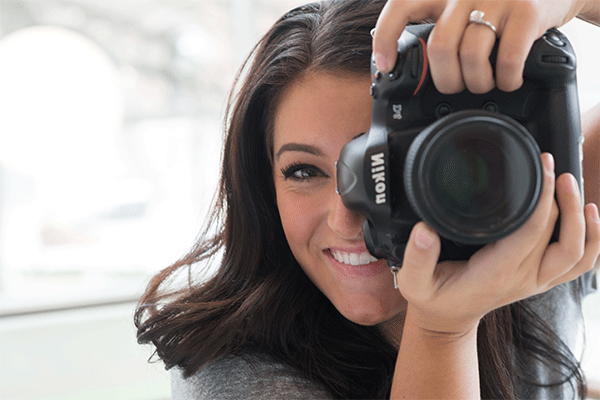 A young female holding a large Nikon camera vertically while looking through the eye piece with the lens the viewer.